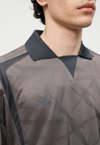 Mand iført en grå sportstrøje med en sort krave og subtile geometriske mønstre, der viser Umbro-logoet på brystet.