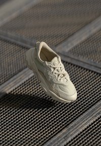 Beige sneakers med texturerat tyg, böjd design och snörning. Noterbara detaljer inkluderar en slät panel på sidan och stoppad krage.