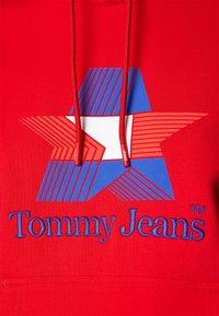 Sweat à capuche rouge avec une étoile géométrique en lignes bleues, blanches et rouges, et "Tommy Jeans" brodé en bleu sous l'étoile.