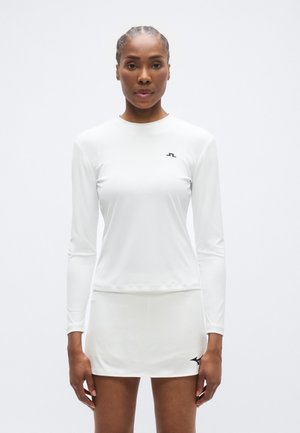 Valge sportlik pika varrukaga pluus ümarate kaeluseäärtega, kombineeritud valge seelikuga. Vasakul rinnal väike must logo ja sile tekstuur.