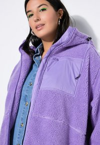 Lila Fleecejacke mit einer strukturierten Oberfläche, ausgestattet mit einer glatten Brusttasche, verstellbarem Kapuze und kombiniert mit einem Jeanshemd darunter.