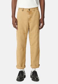 Pantalones cargo color caqui con un corte recto, detalles de costura y puños enrollados. Combinados con zapatos negros y calcetines blancos, sobre un fondo neutral.