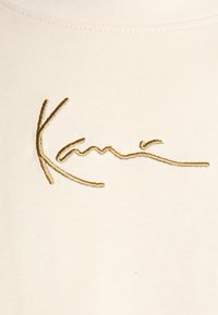 Beige Baumwoll-T-Shirt mit goldbesticktem Unterschriftsdesign, mit glatter Textur und engem Halsauschnitt.