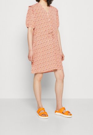 Woman wearing a short-sleeve pink floral dress with a drawstring waist and bright orange sandals with thick white soles.