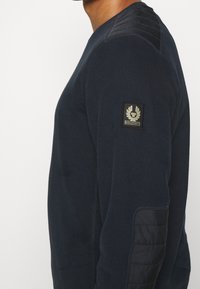 Man med en mörk marinblå långärmad skjorta med kviltade axel- och armbågsfickor samt en Belstaff-logotyp på överarmen.