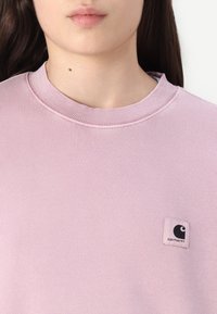 Ljusrosa sweatshirt med ribbad rund halsringning, med en liten svart och vit logotyp på vänster sida. Mjuk, texturerad tyg.