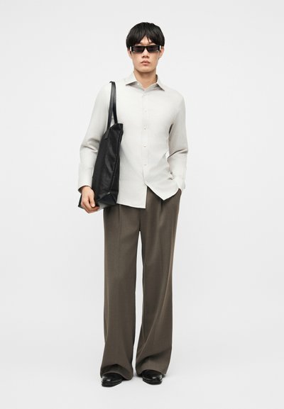 Mann som har på seg en lysebeige skjorte med knapper, brune bukser med vide ben, svarte sko, solbriller og bærer en svart skinnveske på skulderen.