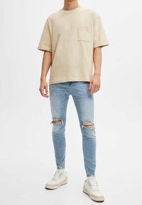 Beige bomullströja med bröstficka, kombinerad med ljusblå denimjeans med rivningar. Modellen har på sig vita sneakers.