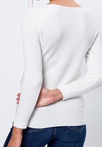 Maglia bianca a maniche lunghe con scollo rotondo e polsini aderenti, che mostra un tessuto testurizzato. Indossata con jeans blu scuro.