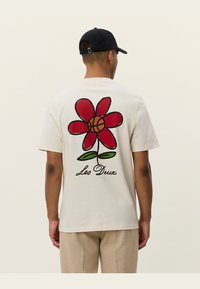 Camiseta de algodón color crema con una gran flor roja en el centro, que tiene un balón de baloncesto en el medio, y el texto "Les Deux" debajo de la flor.