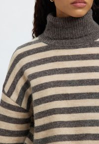 Csíkos pulóver magas bordázott gallérral. Puha, bézs és szürke fonalból készült, vízszintes csíkokkal és laza szabással.