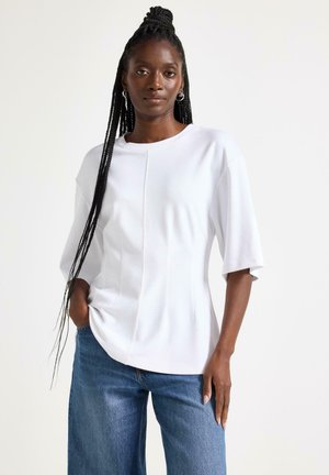 Sort kvinde med langt fletninger iført hvid top med korte ærmer og blå jeans, stående med den ene hånd i lommen mod en ensfarvet baggrund.