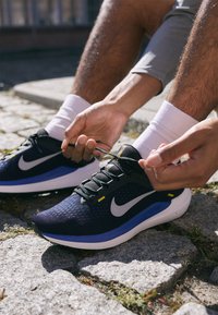 Scarpa atletica nera e blu con tomaia in rete, logo swoosh bianco, lacci testurizzati e dettagli gialli. Indossata con calze bianche su ciottoli.