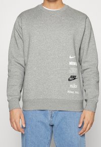 Šedá mikina vyrobená z měkké látky má různé loga Nike v černé a bílé barvě. Má kulatý výstřih a žebrované manžety a lem.