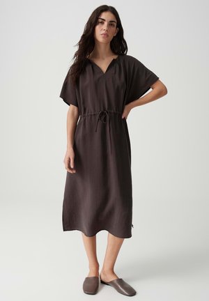 Donna con i capelli lunghi e scuri che indossa un abito marrone scuro, morbido, lungo fino al ginocchio, e scarpe slip-on abbinate, in piedi davanti a uno sfondo chiaro e uniforme.