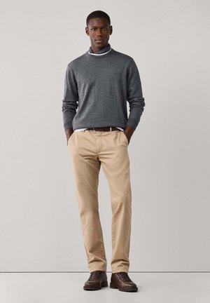 Hombre con jersey gris, bufanda estampada alrededor del cuello, pantalones beige, botas marrones, de pie con las manos en los bolsillos frente a fondo liso.