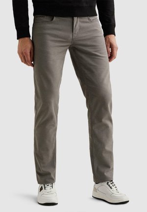 Mand iført grå slim-fit bukser og hvide sneakers, stående mod en ensfarvet baggrund.