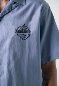 Vaaleansininen, lyhythihainen napillinen paita, jossa on terävä kaulus ja musta kirjailtu logo, jossa on salamaniskuja rinnassa.