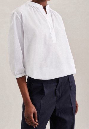 Personne portant un chemisier blanc ample à manches longues avec un petit col en V et un pantalon plissé sombre sur un fond uni.