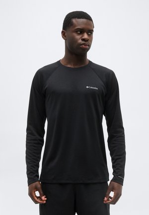 Άνδρας που φοράει μαύρο μακρυμάνικο πουκάμισο Columbia, στέκεται μπροστά σε απλό ανοιχτό φόντο, κοιτάζοντας προς τα αριστερά του.