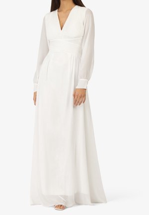 Femme portant une longue robe blanche avec des manches longues transparentes, un profond décolleté en V, une taille cintrée et une jupe fluide, debout devant un fond uni.