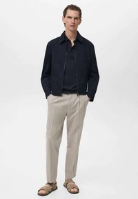 Marine blauwe zip-up jack, lichte stof, gedragen met een donker polo, beige op maat gemaakte broek en bruine sandalen, staande tegen een neutrale achtergrond.