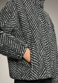 Kurtka z szarego i białego futrzastego materiału w wzór houndstooth, z wysokim kołnierzem i boczną kieszenią, charakteryzująca się luźnym krojem i teksturowaną powierzchnią.