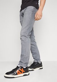 Jeans de mezclilla gris con un corte ajustado, combinados con zapatillas negras que tienen un logo naranja. Los jeans presentan un desgaste sutil y una textura suave.