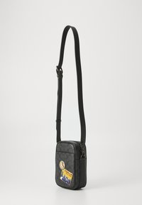 Schwarze Leder-Umhängetasche mit verstellbarem Gurt, ausgestattet mit dekorativen Patches und einer strukturierten Außenseite. Rechteckige Form mit Reißverschluss.