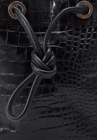 Czarna torba z wytłaczanego krokodylego skóry o połyskującej wykończeniu, z supełkowym zapięciem na sznurku i metalowymi oczkami jako akcenty dekoracyjne.
