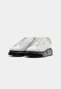 Adidași albi cu un superior din piele netedă, talpă din cauciuc negru, vârf rotund și șireturi plate. Prezintă un logo subtil pe lateral.