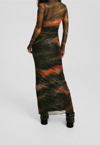 Lång, figurnära klänning i genomskinligt material med mångfärgade abstrakta mönster, främst mörkgrön med röda och gyllene accenter. Försedd med långa ärmar.