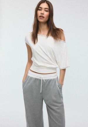 Junge Frau mit langen braunen Haaren, die ein schulterfreies weißes Oberteil und graue Jogginghosen mit Kordelzug trägt, steht mit den Händen in den Taschen.