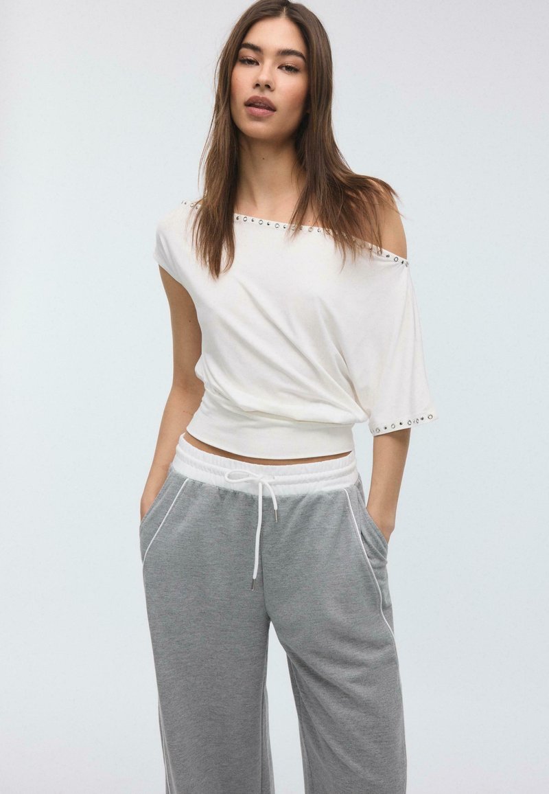 Junge Frau mit langen braunen Haaren, die ein schulterfreies weißes Oberteil und graue Jogginghosen mit Kordelzug trägt, steht mit den Händen in den Taschen.