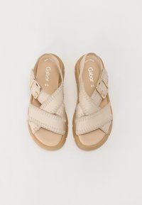 Sandali incrociati beige in pelle, con un motivo decorativo a punto cucito e una fibbia color oro su una suola marrone chiaro.