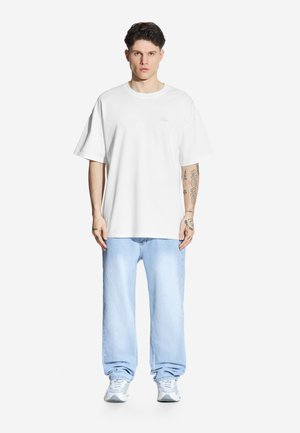 Biała, oversize t-shirt z okrągłym dekoltem, w zestawieniu ze jasnoniebieskimi, prostymi dżinsami. Model nosi szare sneakersy. Prosty i casualowy design.