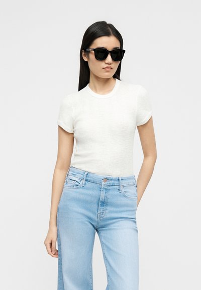 Νεαρή γυναίκα φοράει μαύρα γυαλιά ηλίου, λευκό κοντομάνικο πουκάμισο και ανοιχτόχρωμα τζιν ψηλόμεσα παντελόνια μπροστά σε απλό φόντο.