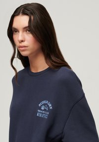 Felpa blu navy con scollatura rotonda e maniche ampie; presenta un logo ricamato blu chiaro "Superdry Beach Club Athletics" sul petto.