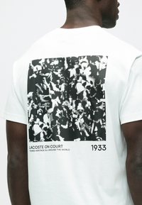 Biała koszulka z krótkim rękawem z czarno-białym nadrukiem graficznym na plecach, z napisem „LACOSTE ON COURT” oraz „1933.”