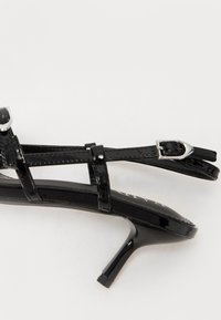 Sorte patentlæder højhælede sandaler med tynde stropper, sølvtonet spænde og spidse tæer. Glat tekstur med en blank overflade.