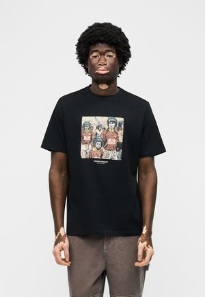 Μαύρος άντρας με λεύκη που φοράει μαύρο T-shirt με εικονογραφημένη ομάδα ανθρώπων με κράνη, με την ετικέτα "Weekend Offender".