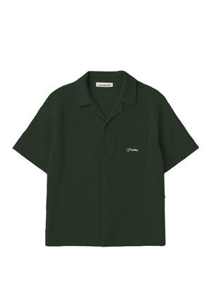 Femme portant une chemise boutonnée à manches courtes vert foncé et une casquette assortie avec un logo NY orange, debout les mains dans les poches sur fond blanc.