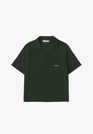 Frau trägt dunkelgrünes kurzärmeliges Hemd mit Knöpfen und passende Kappe mit orangem NY-Logo, steht mit Händen in den Taschen auf weißem Hintergrund.