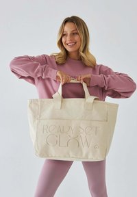 Beżowa torba na ramię z podwójnymi uchwytami, z wytłoczonym tekstem "READY SET GLOW" na przodzie. Gładka tekstura, prostokątny kształt.