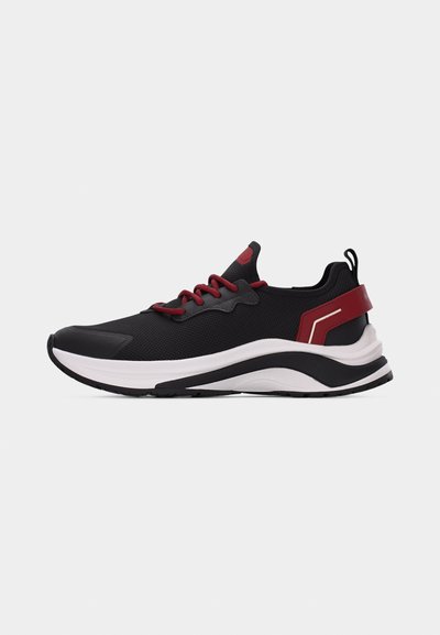 Baskets de sport noires et rouges avec une semelle épaisse blanche et noire, des lacets rouges et une sangle rouge au talon, sur un fond gris clair.