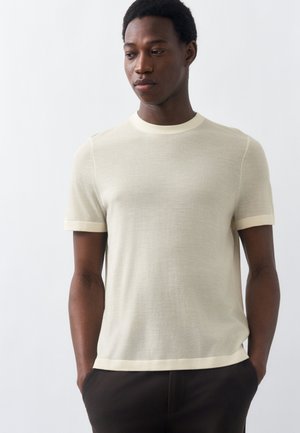 Άνδρας που φοράει ανοιχτό μπεζ κοντομάνικη πλεκτή μπλούζα και σκούρο παντελόνι, στέκεται με τα χέρια στις τσέπες, κοιτάζοντας ελαφρώς προς τα κάτω και πλάγια.