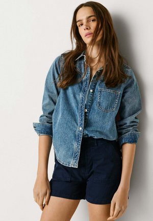 Mladá žena s dlhými hnedými vlasmi, ktorá má na sebe ohrnutú rifľovú košeľu a tmavomodré šortky, stojaca pri obyčajnej bielej stene.