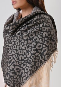 Wrap leopardato realizzato in morbido tessuto, con un design grigio scuro e beige e bordi frangiati, elegantemente drappeggiato sulle spalle della modella.
