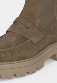 Bottine mocassin en daim marron avec une semelle en caoutchouc beige épaisse et un panneau latéral élastique, présentant des coutures détaillées et un design moc toe.