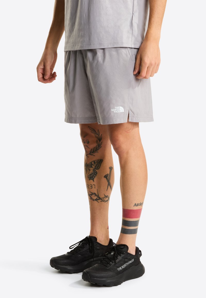 Hellgraue Shorts mit einem dezenten strukturierten Muster, ausgestattet mit einem elastischen Bund und einem weißen Logo. Schwarze Sportschuhe mit Schnürsenkeln und Polsterung.